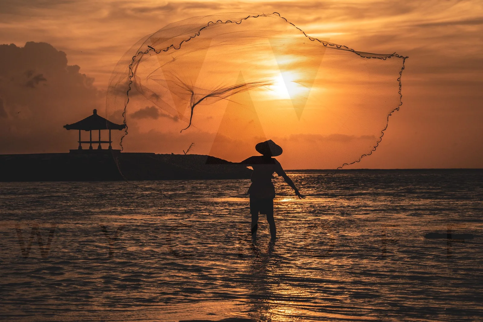 A fisher casts a large net into the sea at sunset