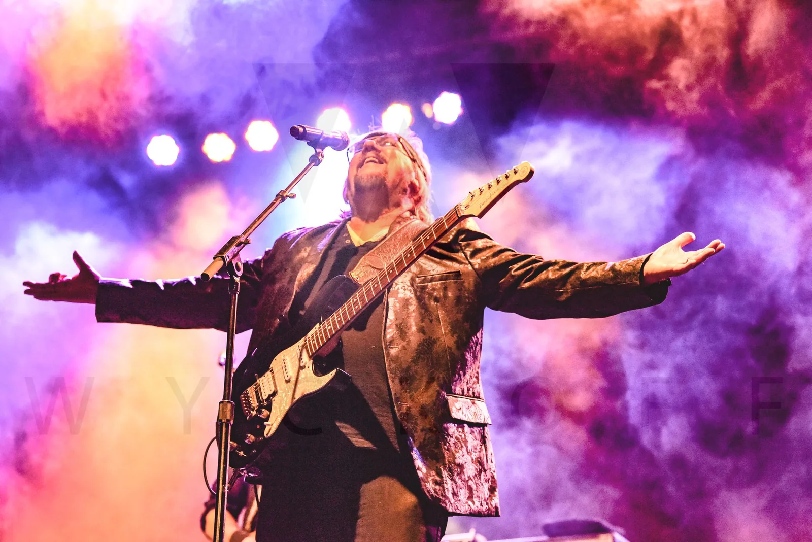 A guitarist spreads his arms beneath colorful stage lights and smoke